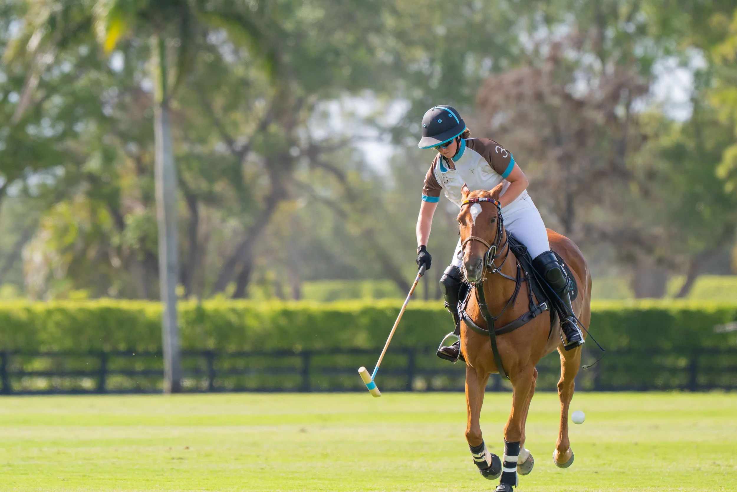 A woman riding a horse on a golf course, wearing sports attire and a helmet, hitting a golf ball.