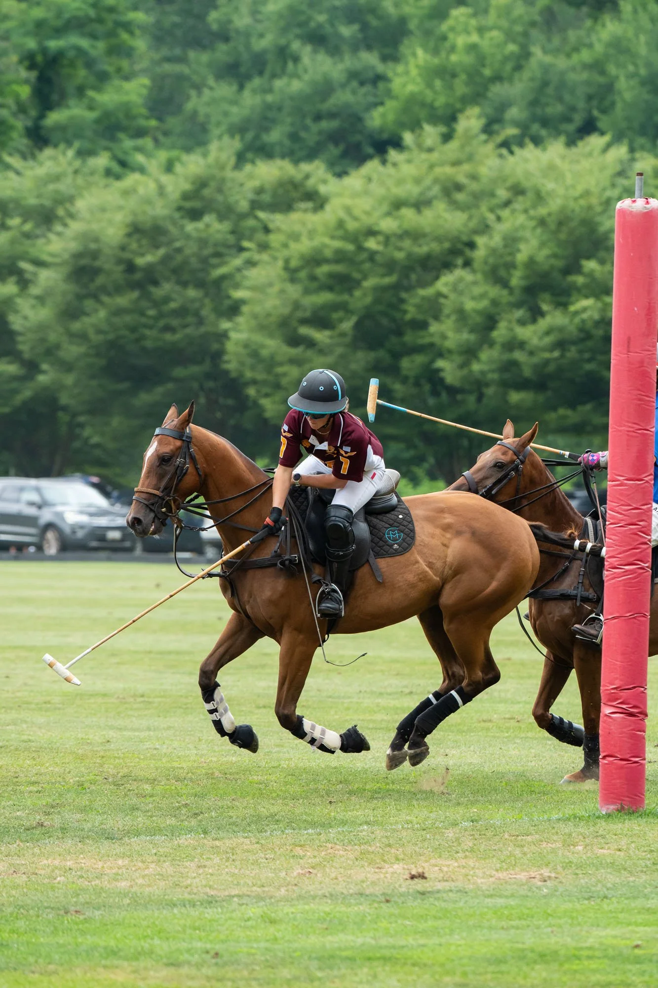 A person wearing a helmet and sports attire riding a horse in a polo match, with another horse and player partially visible, on a grassy field with a backdrop of trees and parked cars.