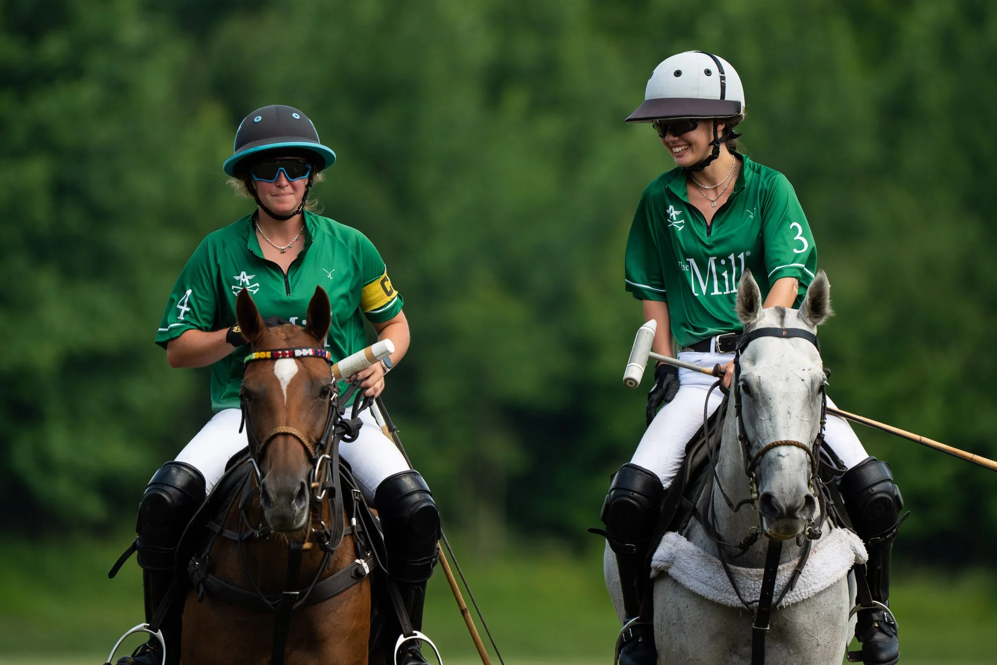 Two women wearing green polo shirts, helmets, and sunglasses riding horses on a lush green field, smiling and enjoying a polo game.
