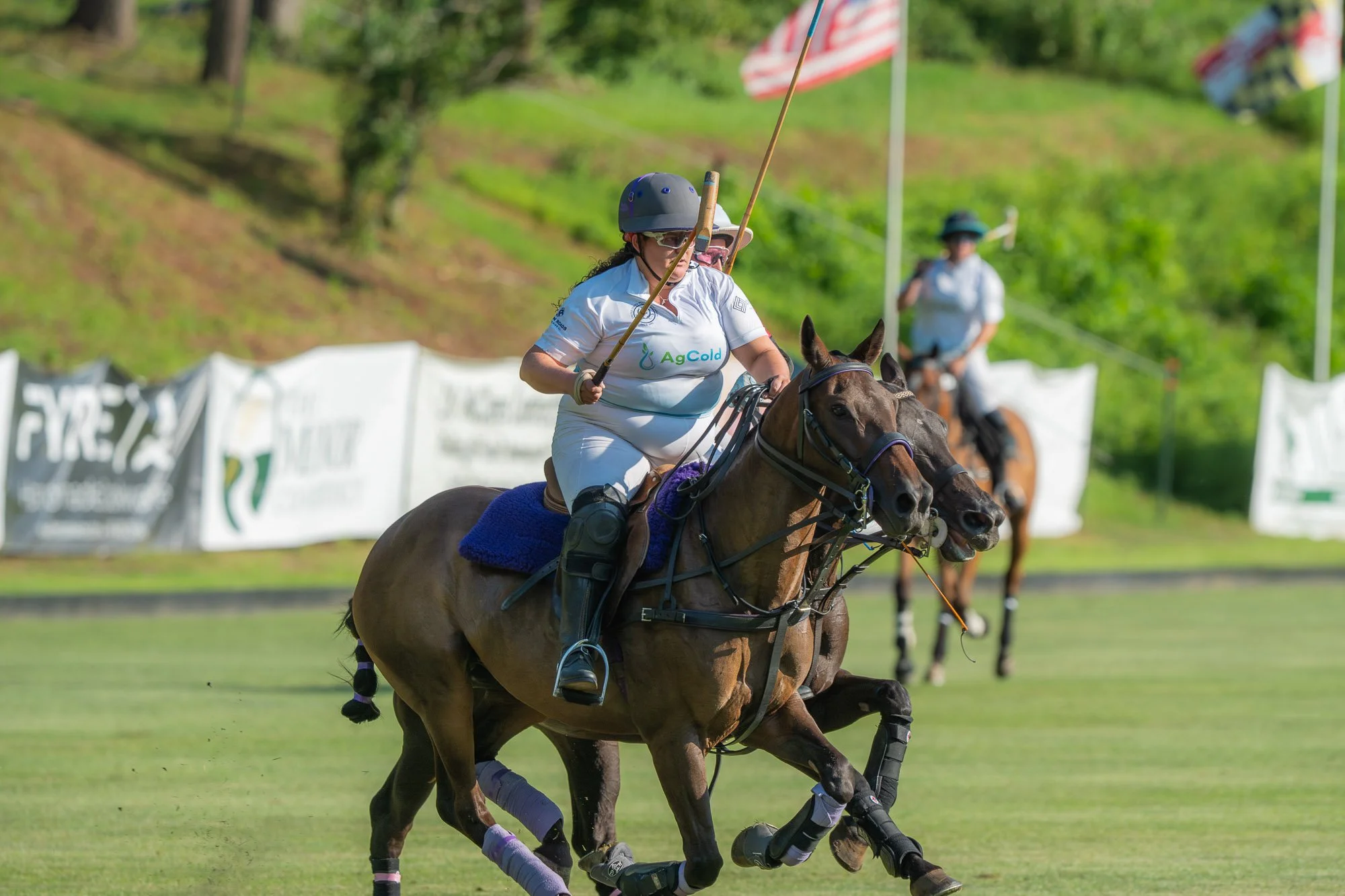 Two women riding horses in a polo match. The woman in front wears a helmet and sunglasses, holds a mallet, and is dressed in a white jersey with the word 'AgCold' on it. The second woman is slightly behind, wearing a hat, sunglasses, and a white jers