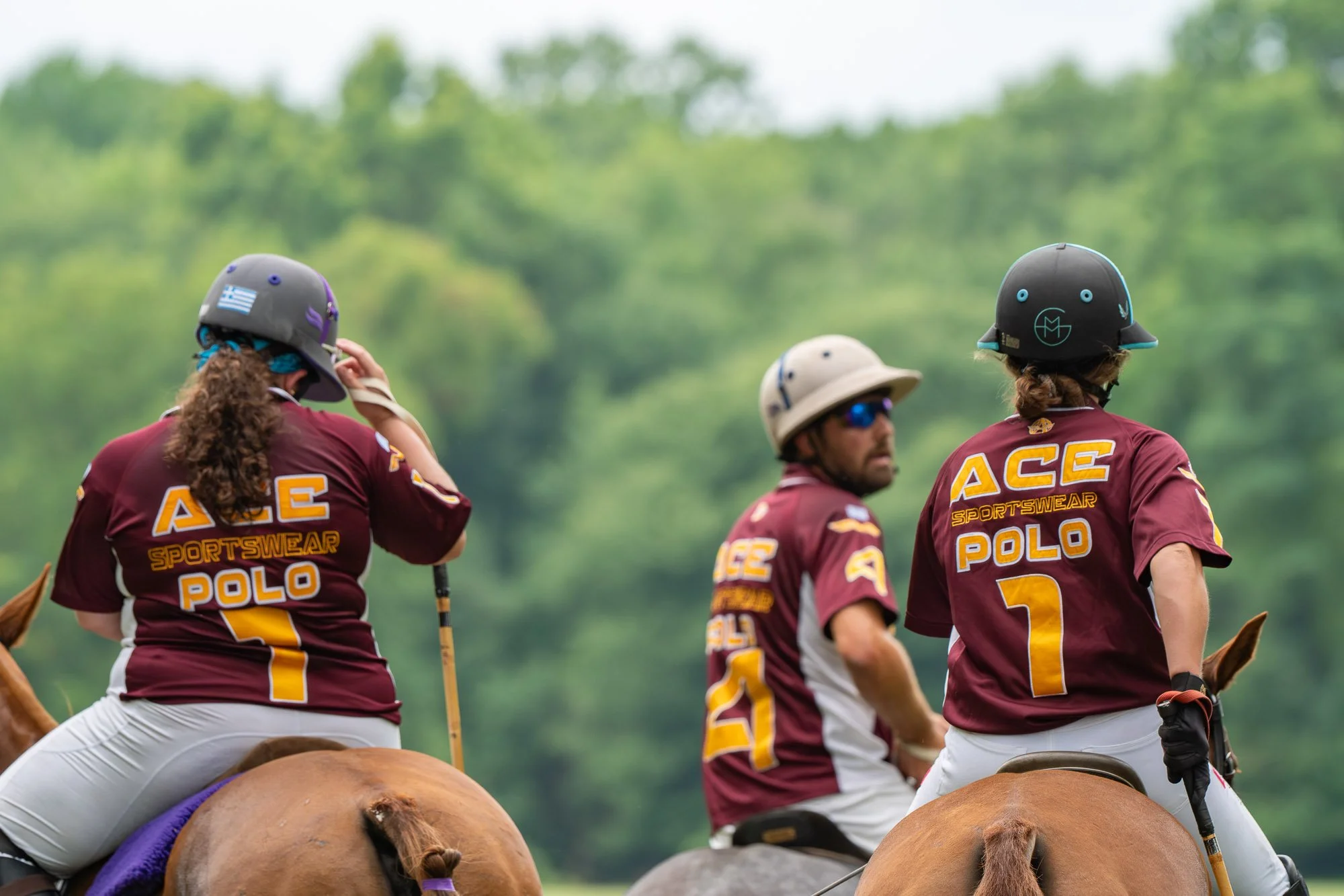 A group of polo players riding horses on a lush green field during daytime. They are wearing matching maroon uniforms with yellow lettering and numbers, and protective helmets. The scene captures the athletes in action, with trees blurred in the back