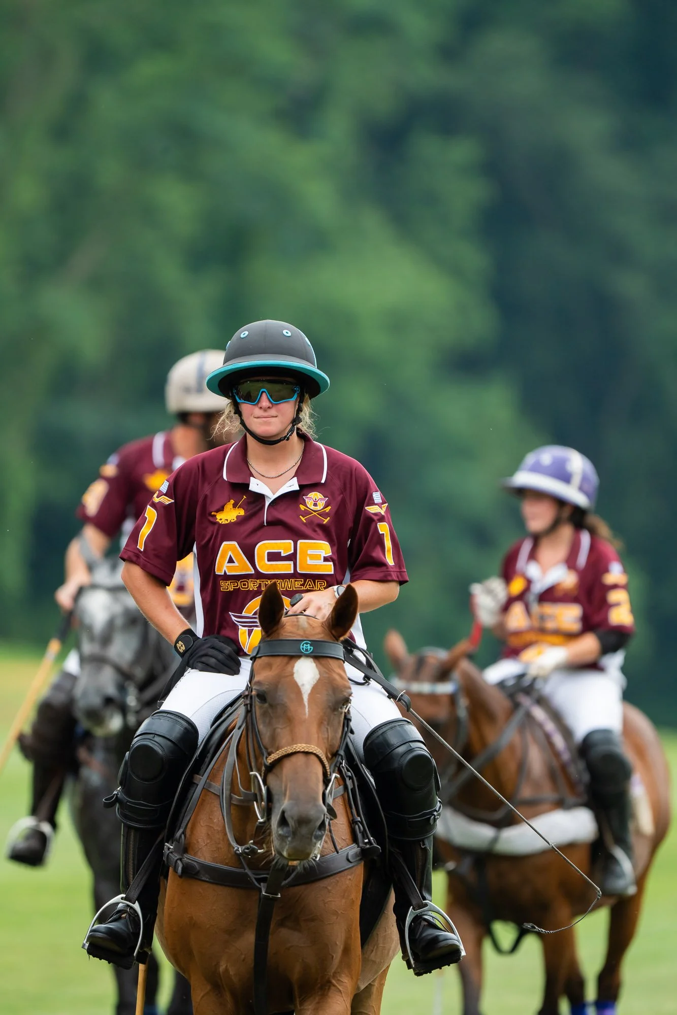 A woman wearing a maroon polo shirt, sunglasses, and a helmet rides a horse in a lush green field with other women on horseback in the background.
