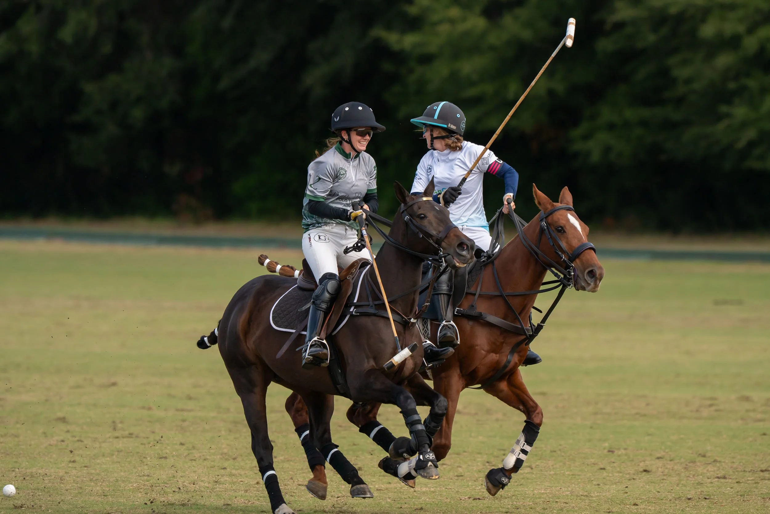 Two women playing polo on horseback in a grassy field, holding mallets and wearing helmets.