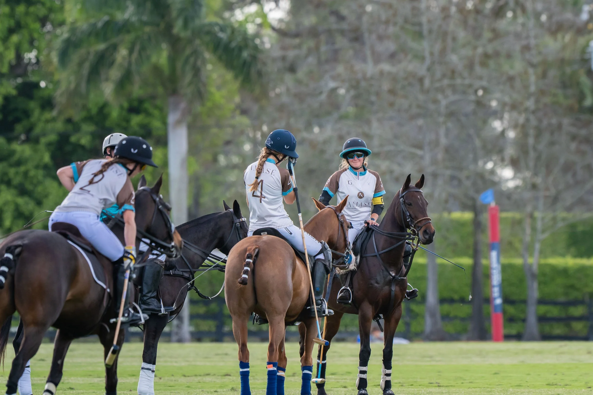 A group of polo players on horseback wearing helmets and jerseys, smiling and holding mallets, on a polo field with trees in the background.