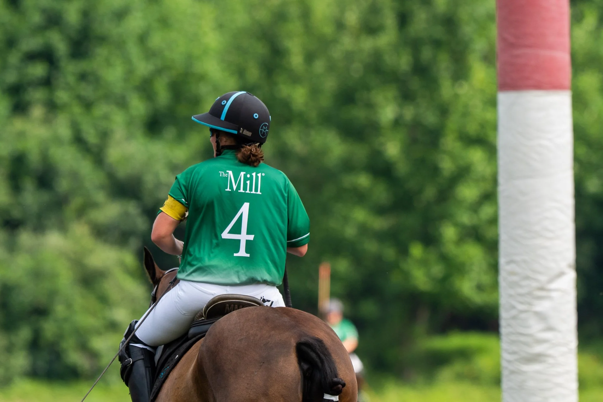 A polo player wearing a green jersey with the number 4 on the back, a black riding helmet, and white pants, riding a brown horse during a game. A red and white goalpost is visible on the right, with lush green trees in the background.
