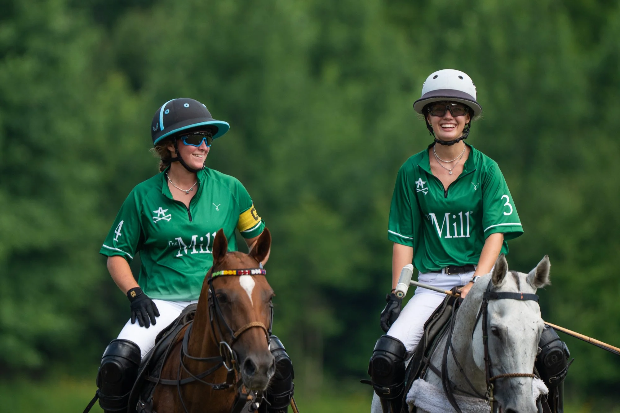 Two women riding horses on a polo field, both wearing green jerseys, helmets, and sunglasses, smiling and enjoying the game.