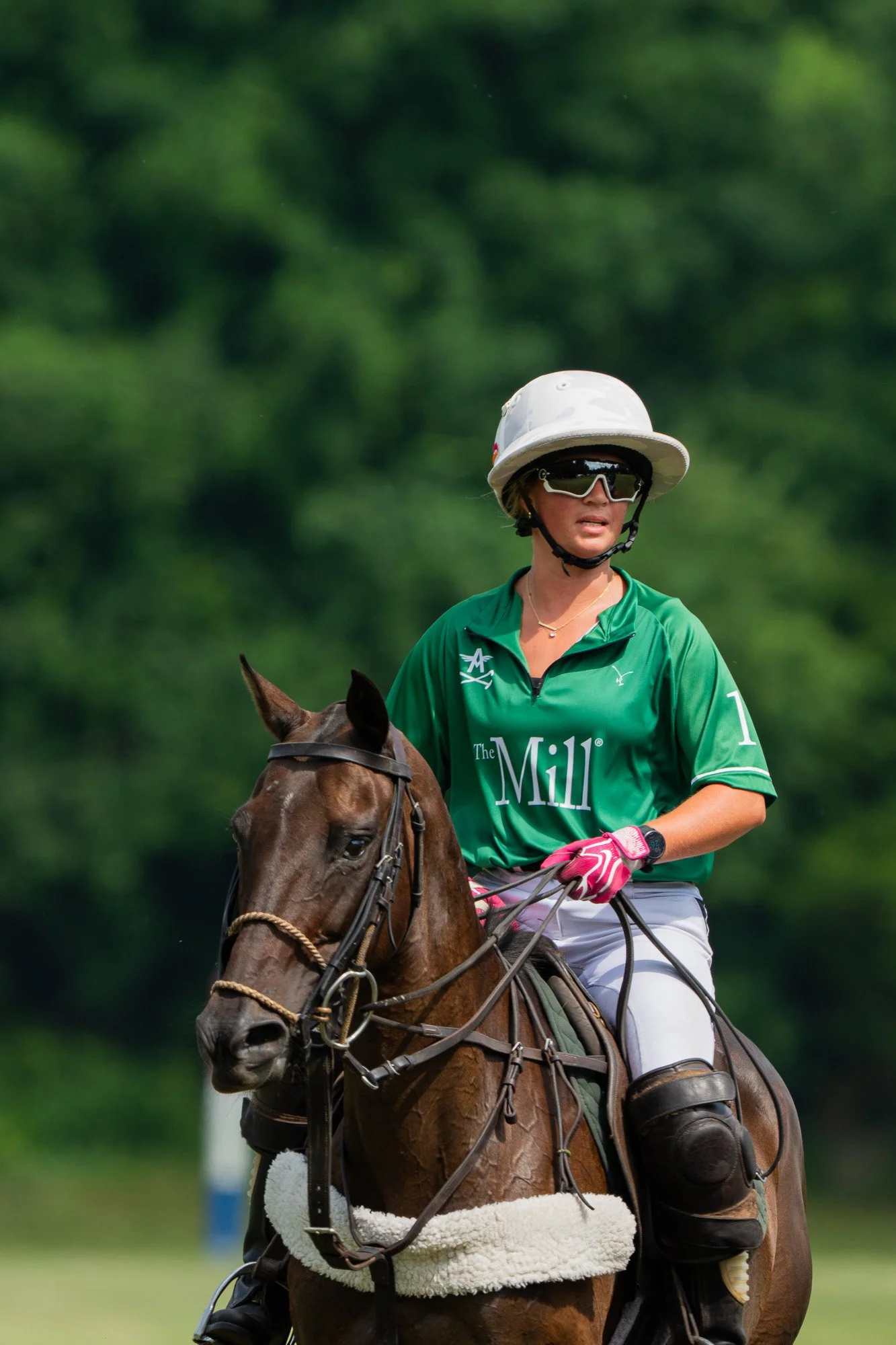 A woman in a green sports shirt and white helmet riding a brown horse during an outdoor event, with a blurred green forest background.