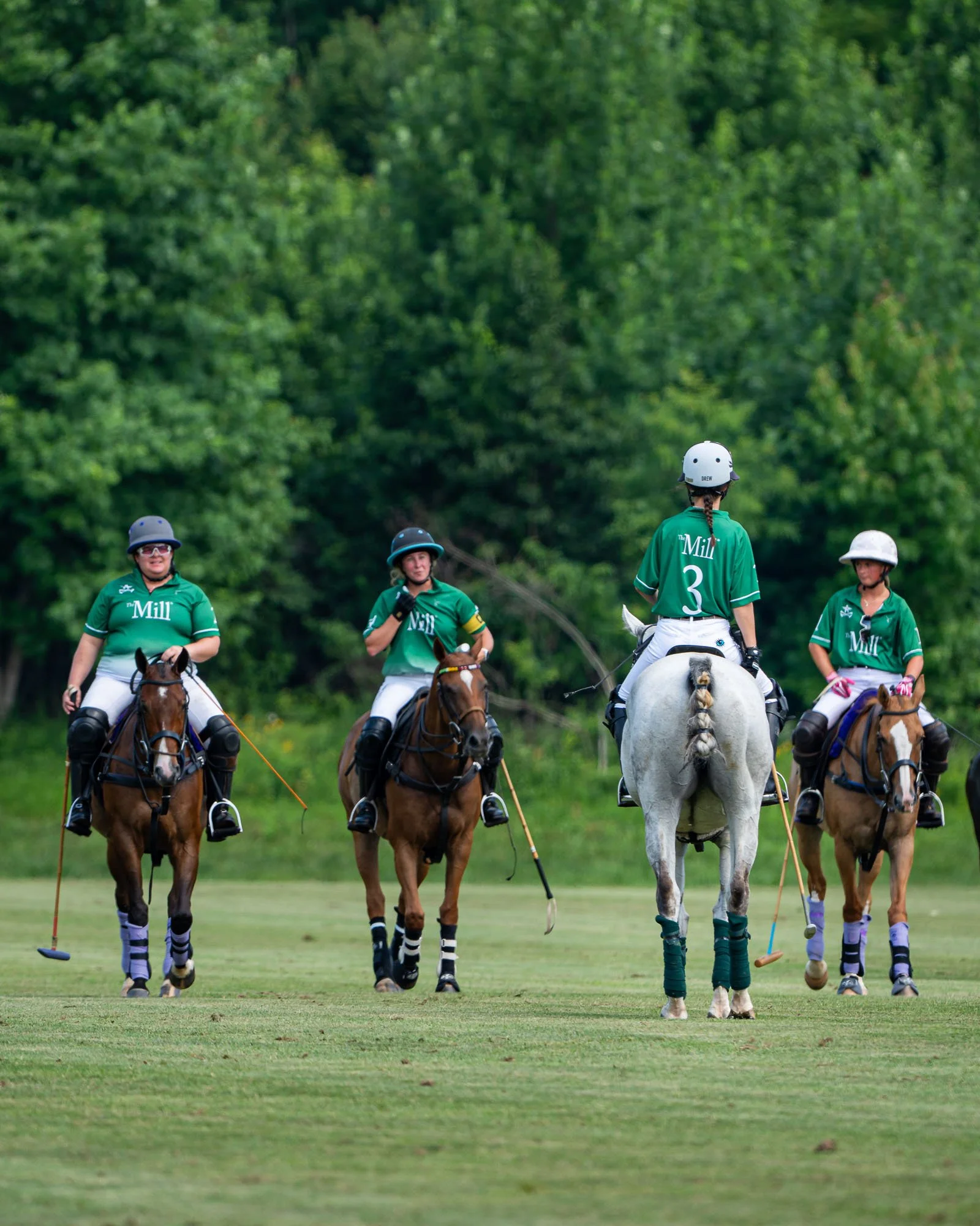 Women riding horses and holding polo sticks on a polo field with a lush green background.