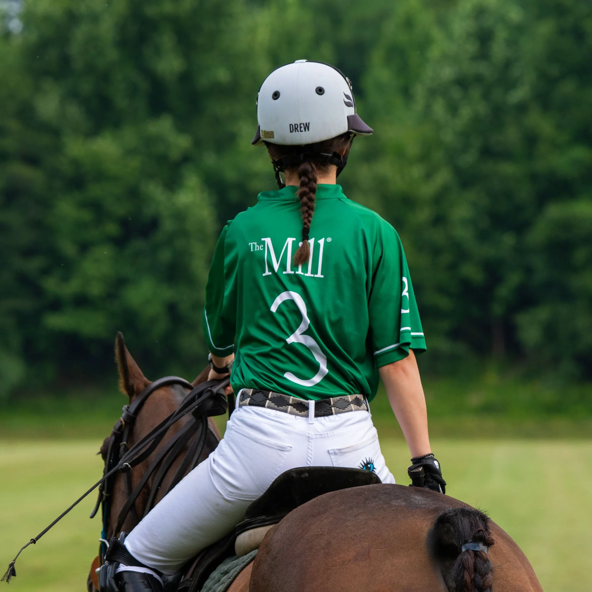Back view of a female horseback rider wearing a white helmet with 'DREW' written on the back, green jacket, and white riding pants, riding a brown horse with a braided mane in a lush green outdoor setting.