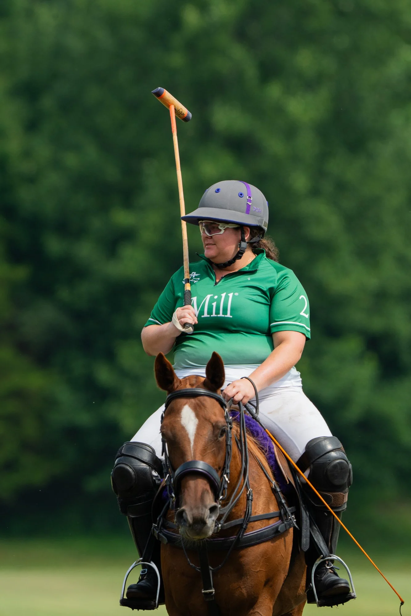 A woman riding a horse outdoors, wearing a helmet and sunglasses, holding a mallet, with a green background.