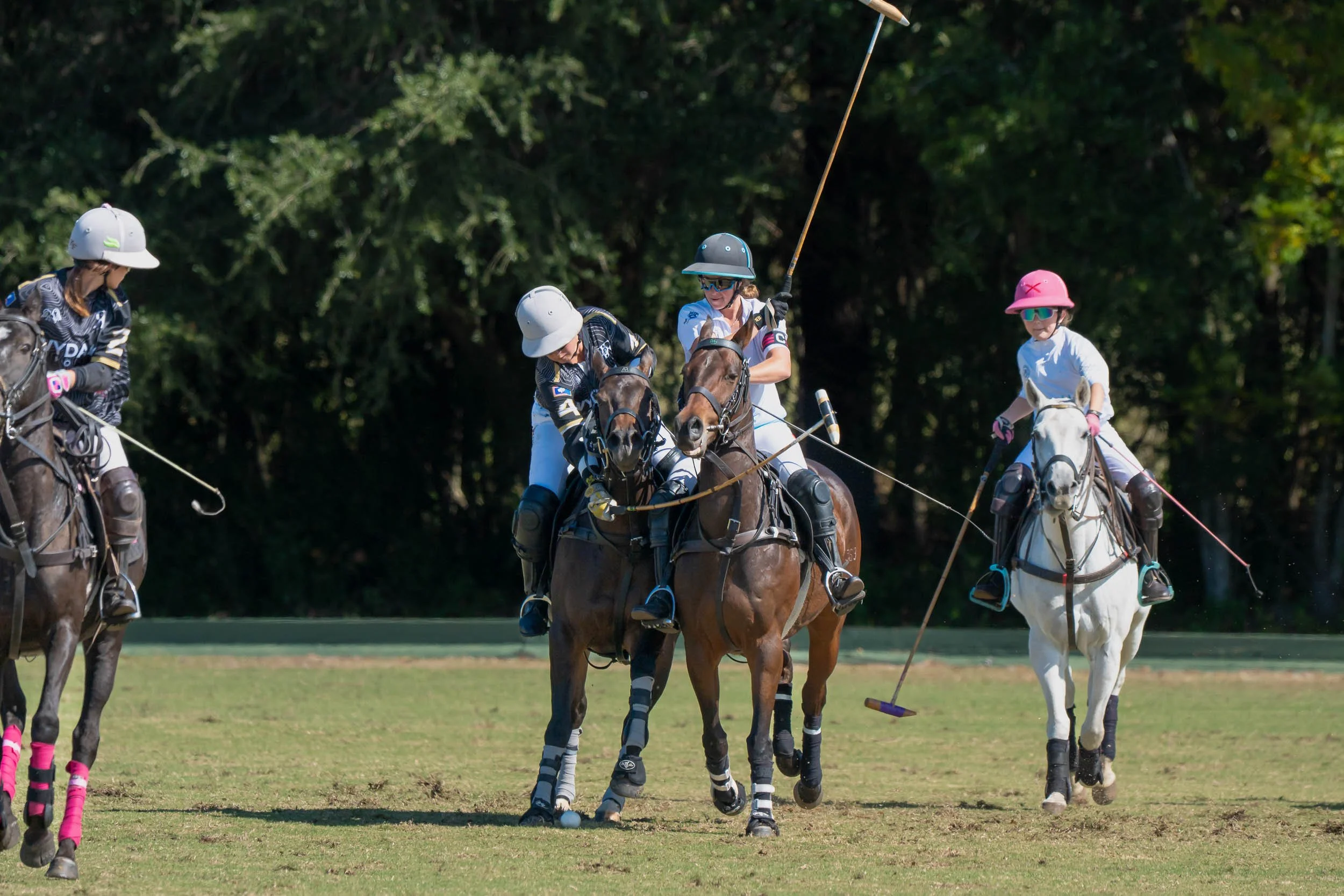 Polo players wearing helmets and protective gear ride horses across a grassy field.