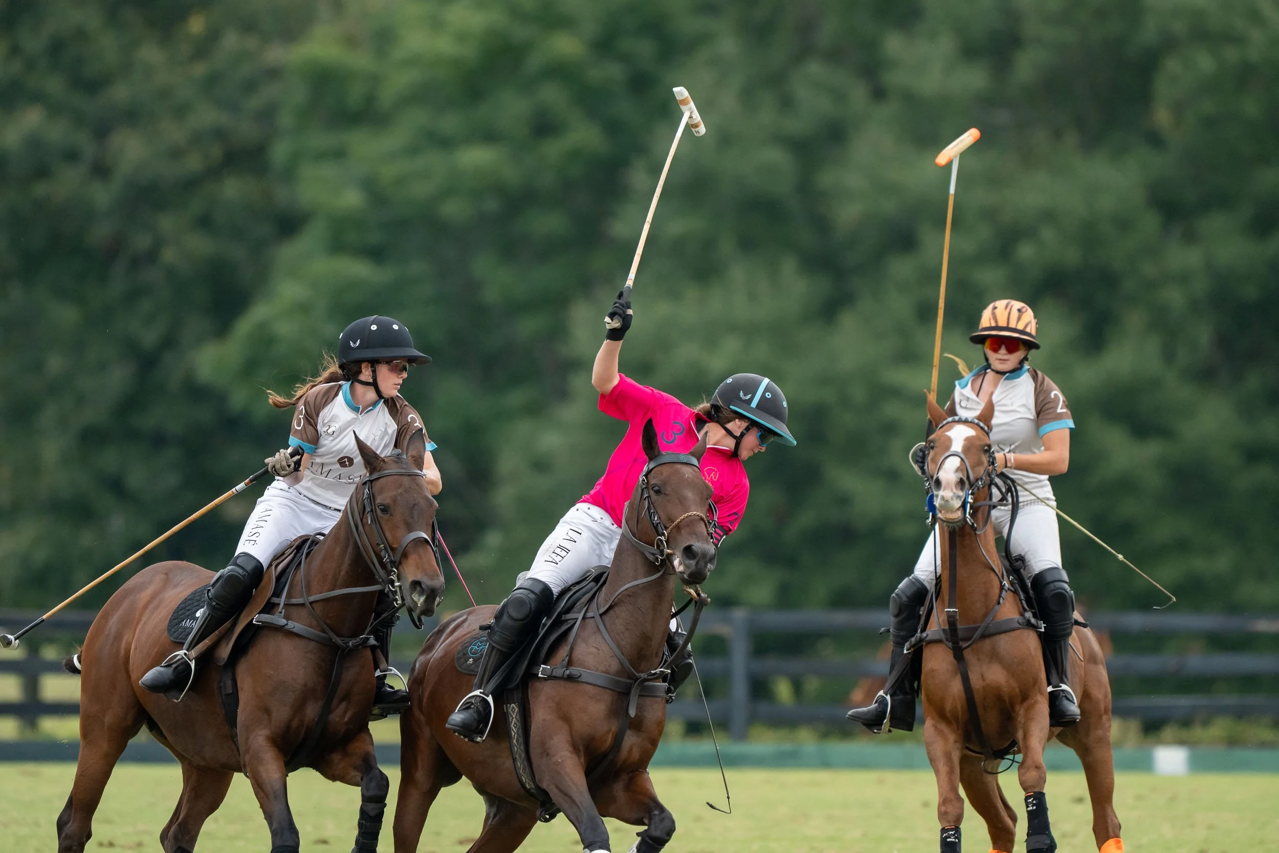 Three female polo players on horseback competing on a grass field, swinging mallets.