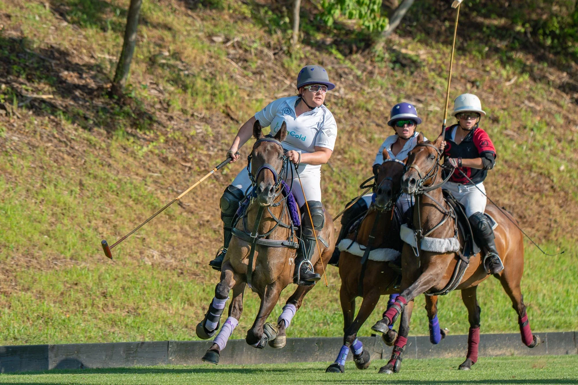 Three women riding horses, participating in a polo match on a grassy field, wearing helmets and protective gear.