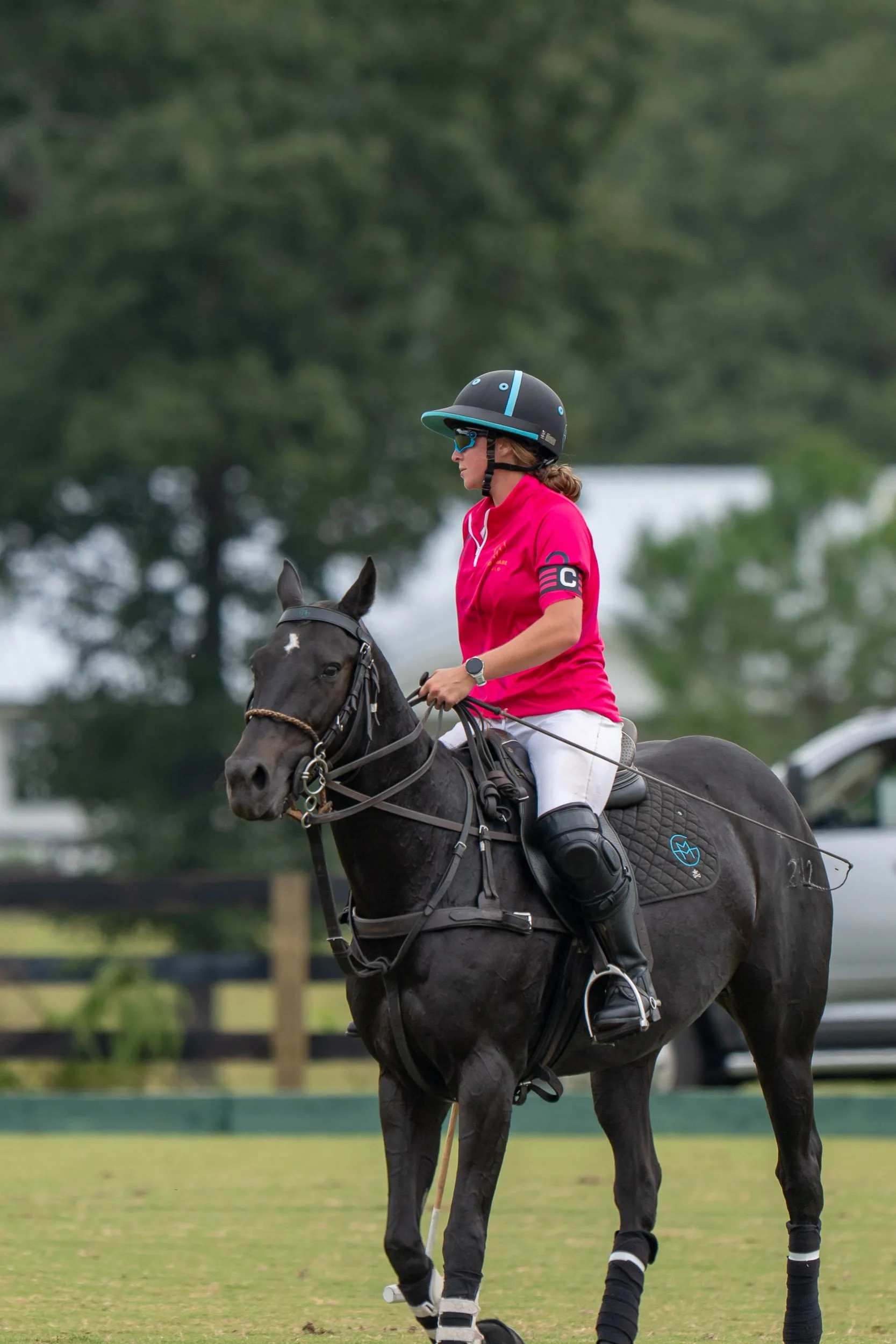 Polo player in a pink jersey and helmet riding a black horse on a grass field