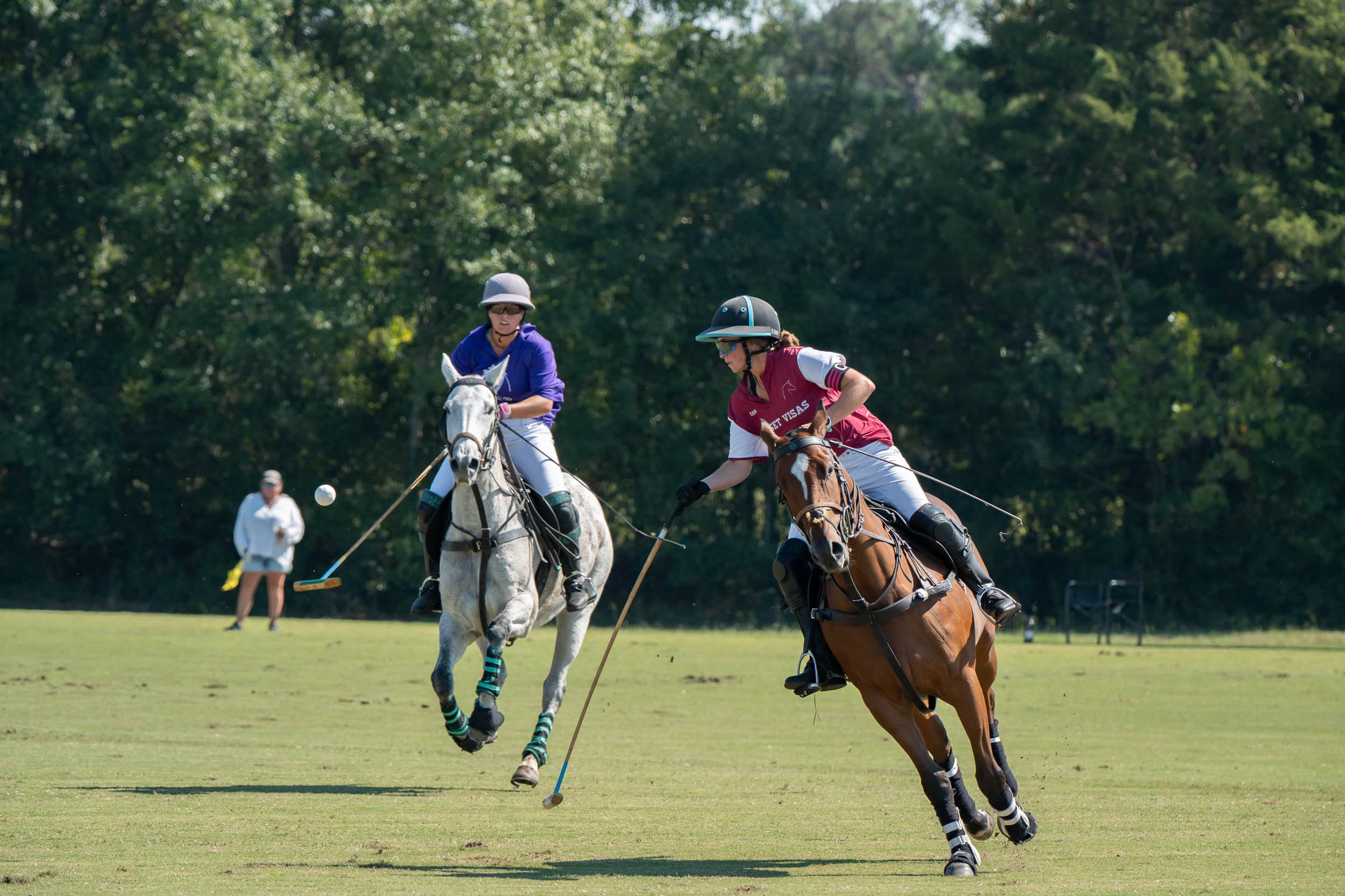 Two horseback riders playing polo on a grassy field, with a polo ball and spectator in the background