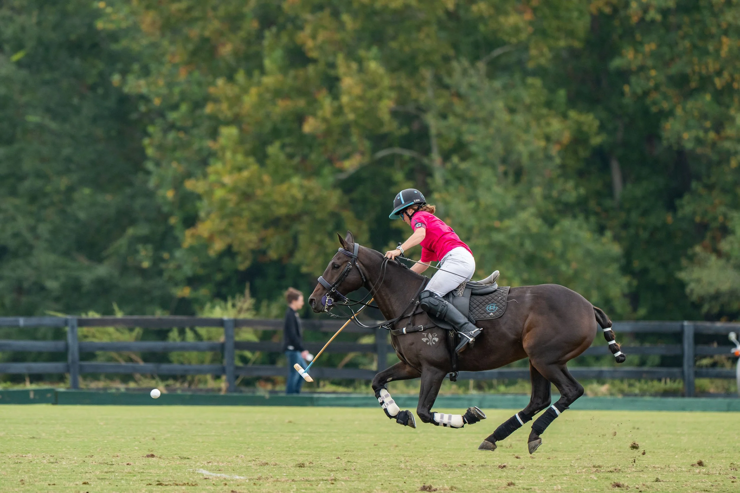 Polo player in a pink shirt riding a galloping horse and swinging a mallet toward the ball on a grassy field