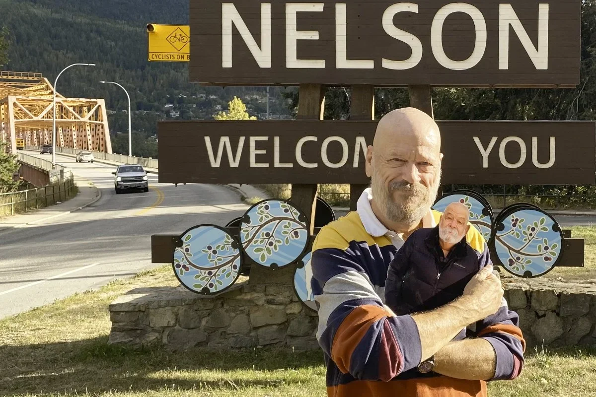 A man with a beard and colorful striped shirt standing in front of a sign that says 'Nelson Welcome You,' with a small cutout of his face in the sign. The sign is on a stone base and there is a bridge and cars in the background.