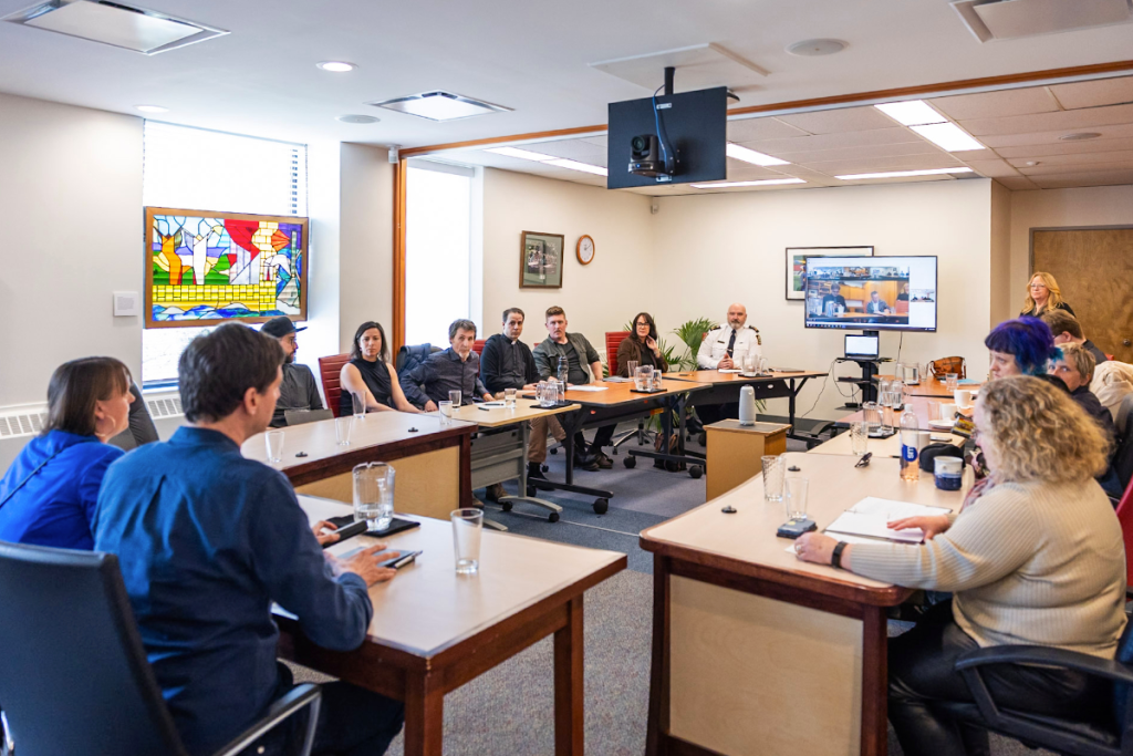 Business meeting in a conference room with people seated around tables, a woman speaking at the front, a large screen displaying a video call, and artwork on the walls.