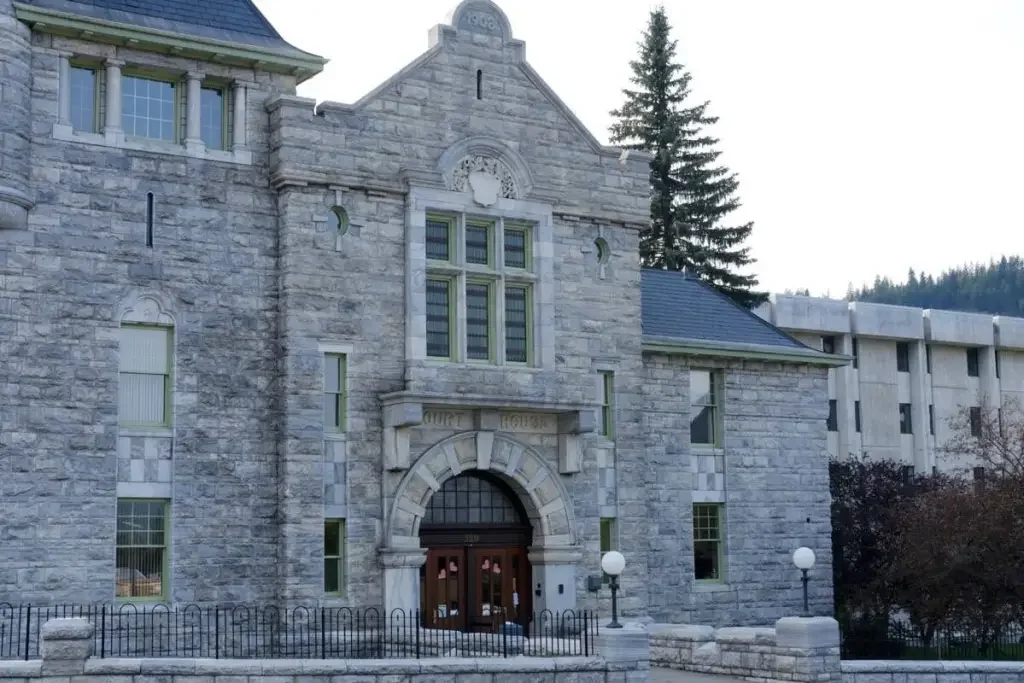 Historic stone building with arched entrance and large windows, surrounded by a small fence and outdoor lamps, with a backdrop of trees and a modern building.