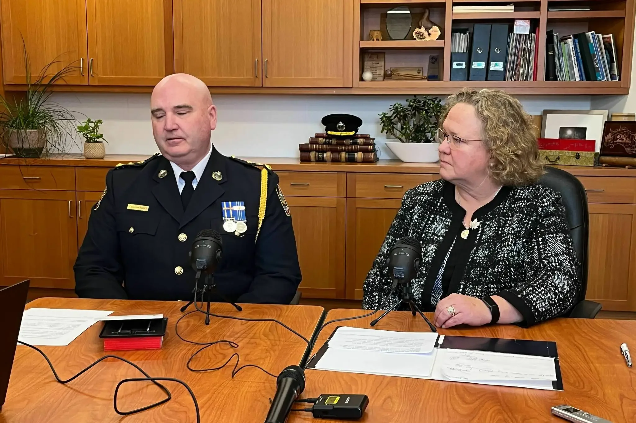 A man in a police uniform and a woman in glasses sitting at a conference table with microphones, papers, and a laptop, in a room with wooden cabinets and bookshelves behind them.