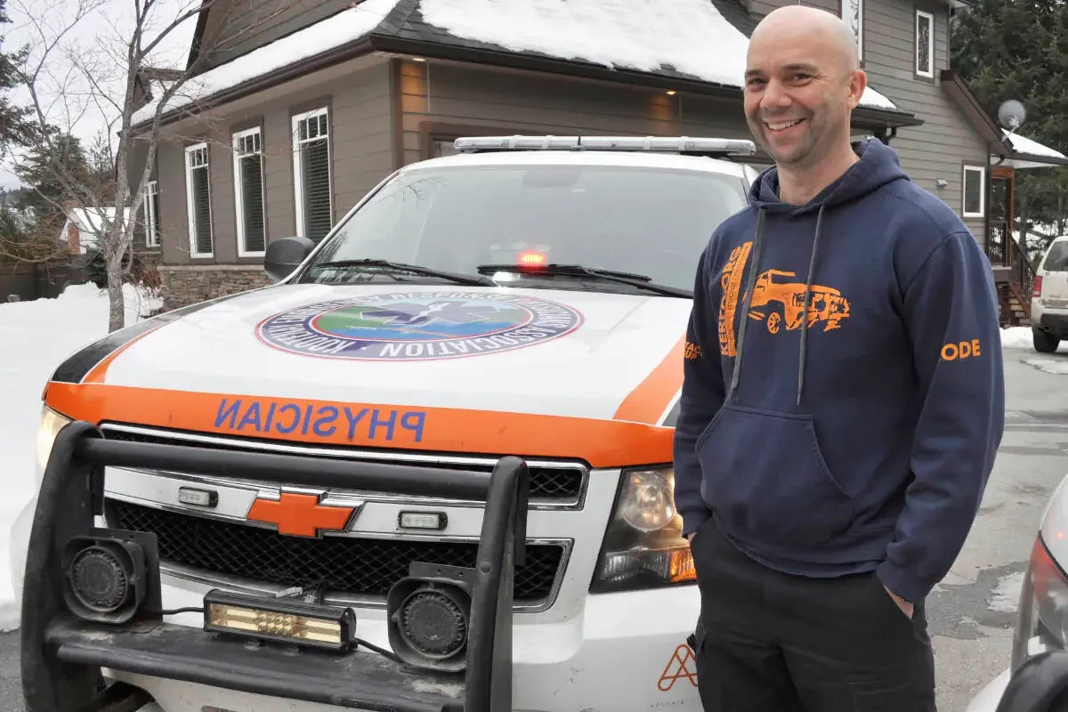 A man smiling and standing next to a white rescue vehicle with emergency lights on a snowy day in front of a house. The vehicle has 'Rescue' written upside down and an emergency insignia on the hood.