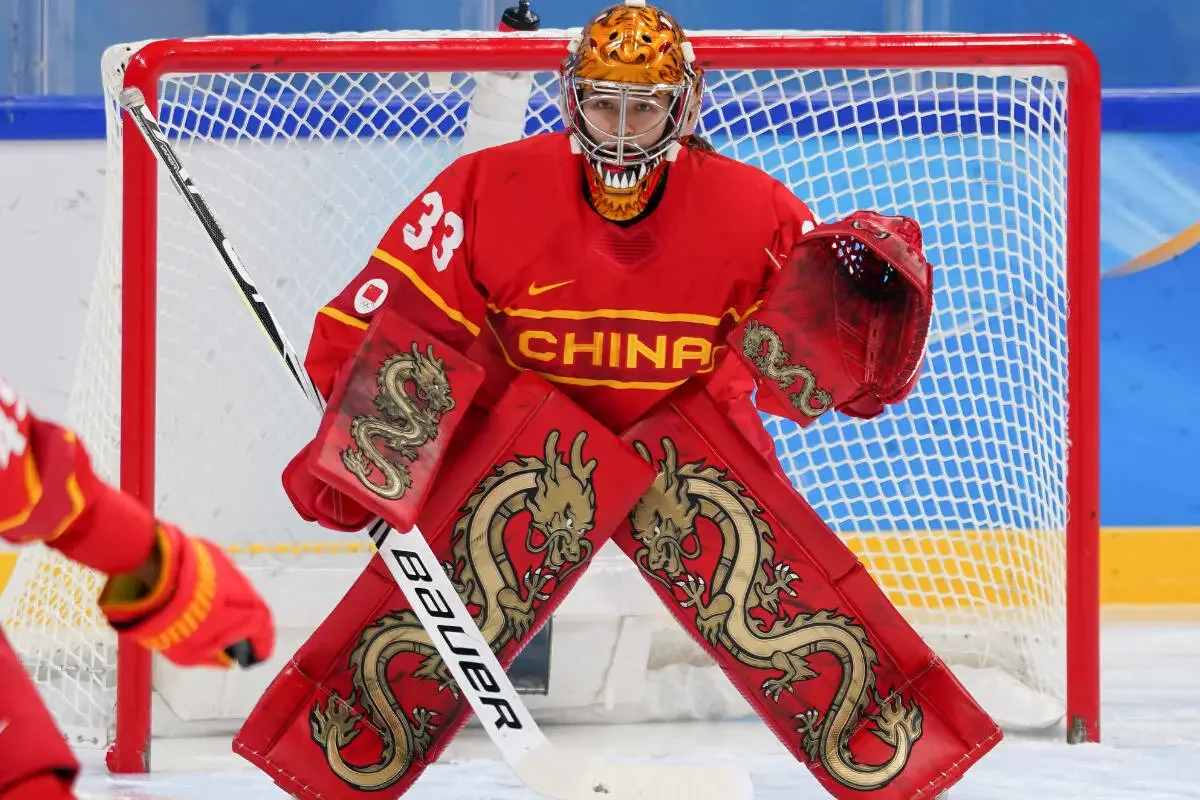 Hockey goalie wearing a red China team uniform with dragon designs, yellow and red helmet, standing in front of goal during a game.