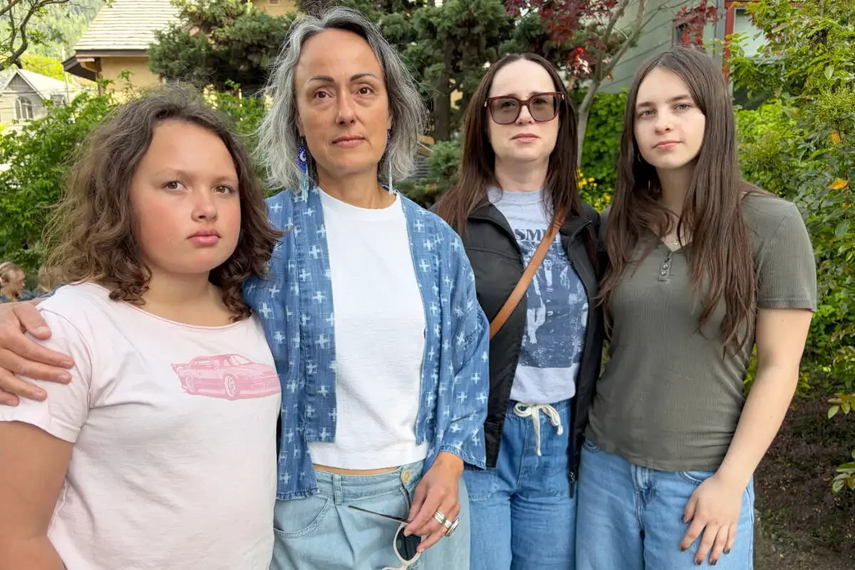 Group of four women outdoors in a garden or park, standing side by side and looking at the camera. The woman on the left has curly brown hair and a light pink shirt. The woman second from the left has gray hair, wearing a white top with a blue patterned jacket. The woman second from the right has long brown hair, glasses, and wears a black vest over a graphic T-shirt. The woman on the right has long dark brown hair and is dressed in a gray T-shirt. Background includes trees and shrubs.