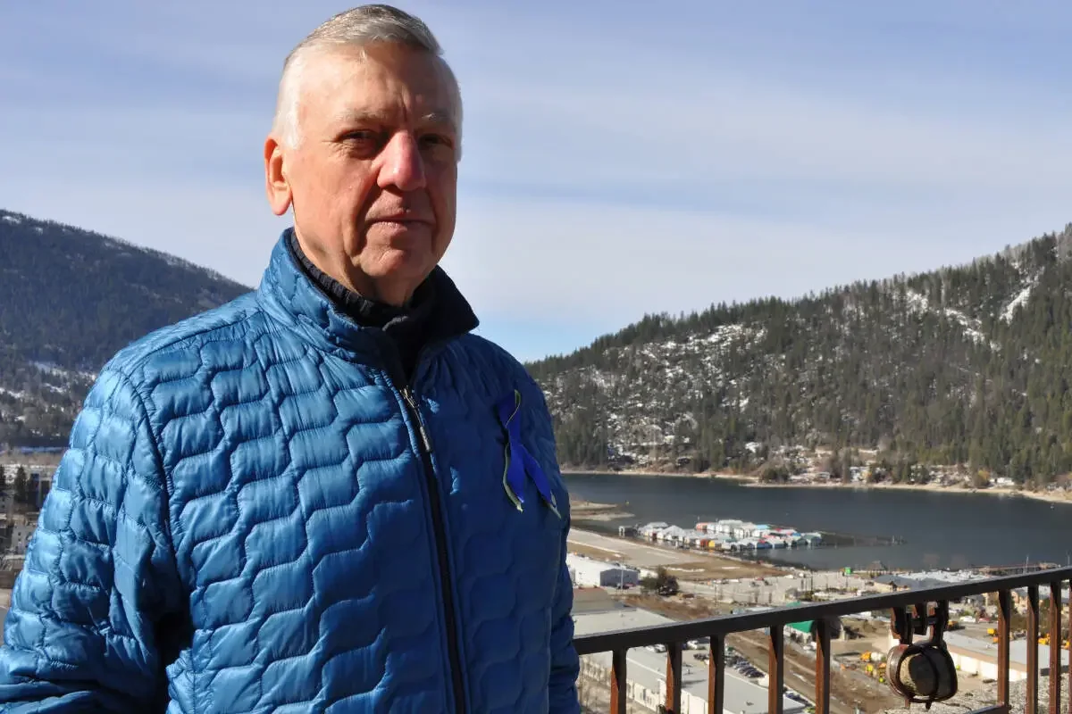 A middle-aged man in a blue quilted jacket standing outdoors on a balcony with a landscape of mountains, a lake, and a small town or industrial area in the background.