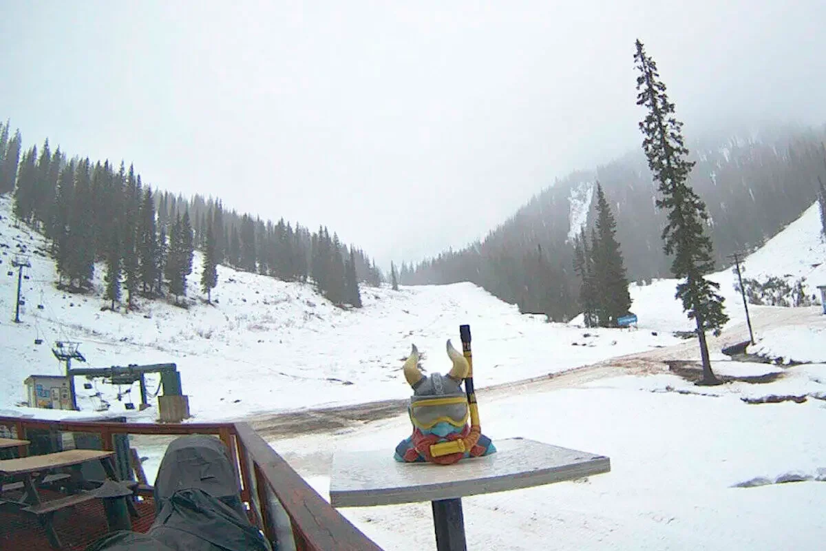 Snow-covered mountain ski resort with ski lift, pine trees, and cloudy sky. A colorful ski figurine with horns and goggles is on a table in the foreground.