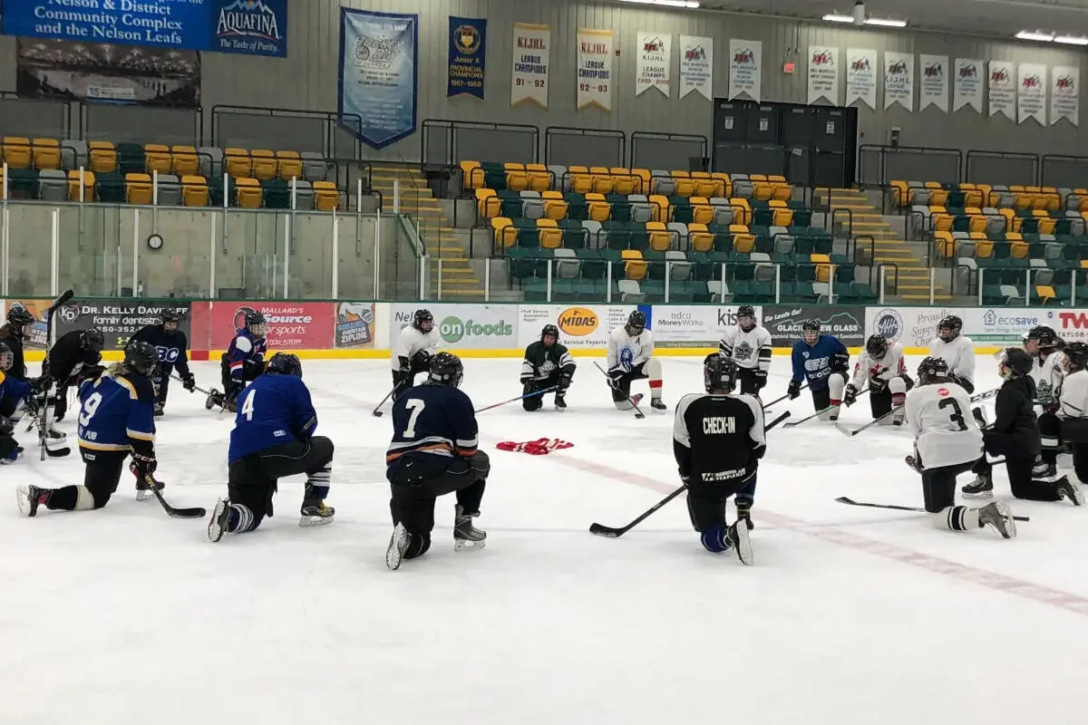 Hockey team kneeling on ice rink during practice or meeting, with players wearing jerseys and helmets, and coach or trainer standing in front. Empty spectator seats and banners in the background.