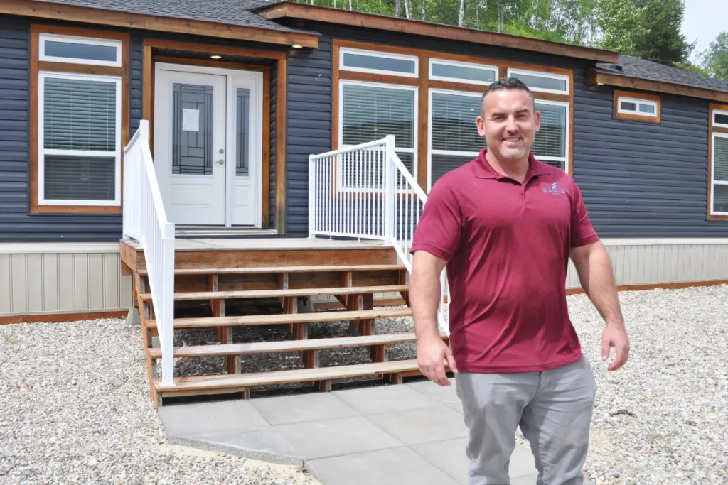 Man standing in front of a house with a porch and stairs, wearing a red polo shirt and gray pants.