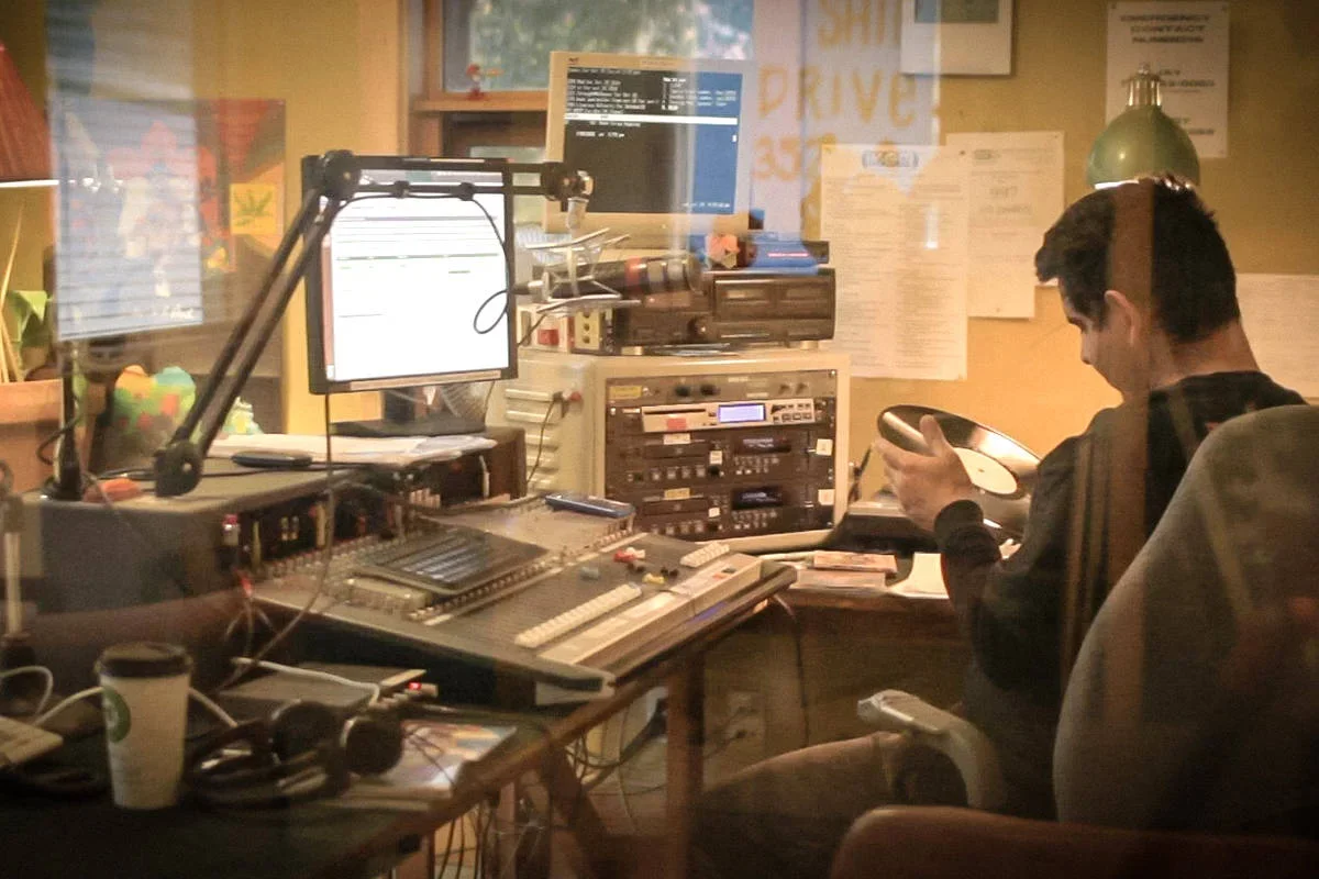 A radio station studio with a man sitting at a desk looking at a phone, surrounded by audio equipment, computer monitors, and papers on the wall.