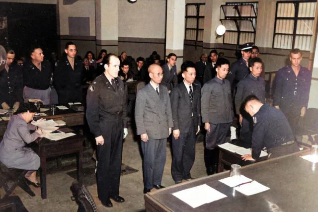A courtroom scene with a group of people standing before a judge. Several individuals are dressed in suits and uniforms, with some taking notes at desks.