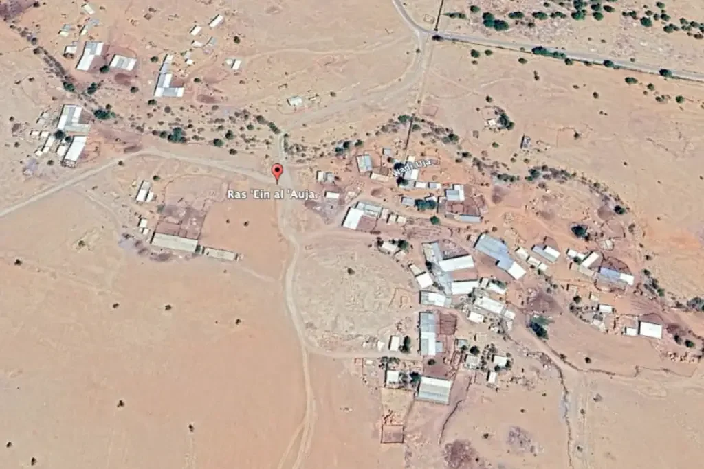 Satellite view of a small, dusty desert village with scattered buildings and dirt roads, labeled Ras 'Ein al 'Auja.