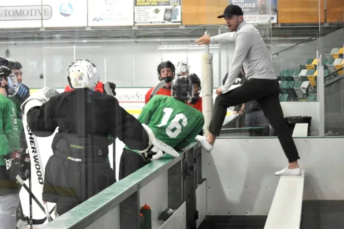 A hockey coach or trainer in a gray shirt and black pants is standing on the sideline of an ice rink, leaning over a ledge, while a hockey player in a green jersey with the number 16 is leaning on the ledge for support after a game or practice. Several other players in red and black gear are watching, with some wearing helmets and face masks.