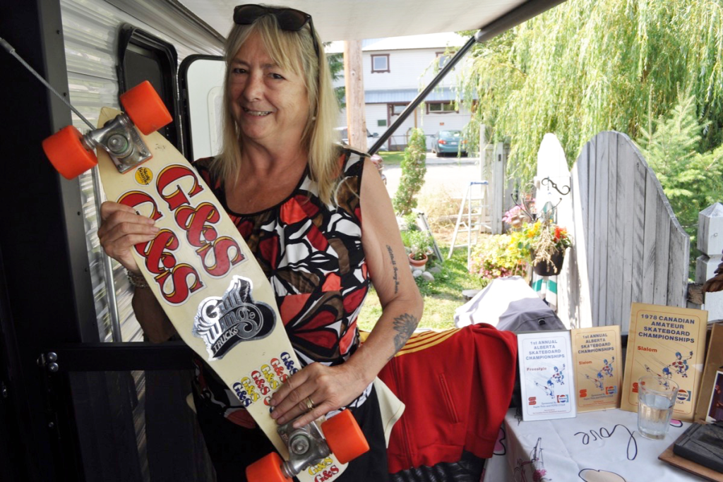 A woman with blonde hair wearing sunglasses on her head and a sleeveless top, smiling while holding a skateboard with orange wheels and a yellow design, standing next to a table with skateboarding event flyers and a glass of water, outdoors in a yard with trees and a fence.