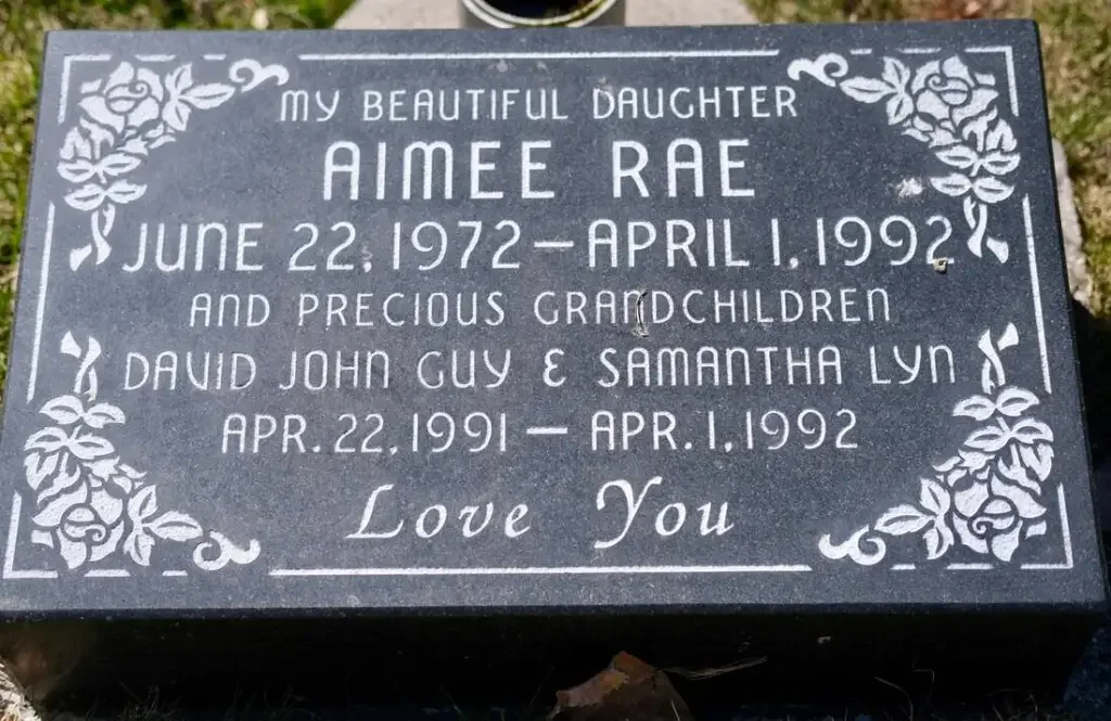 engraved memorial plaque for Aimee Rae, born June 22, 1972, died April 1, 1992, and her grandparents David John Guy and Samantha Lyn, born April 22, 1991, died April 1, 1992, with floral decorations in the corners and the words "Love You".