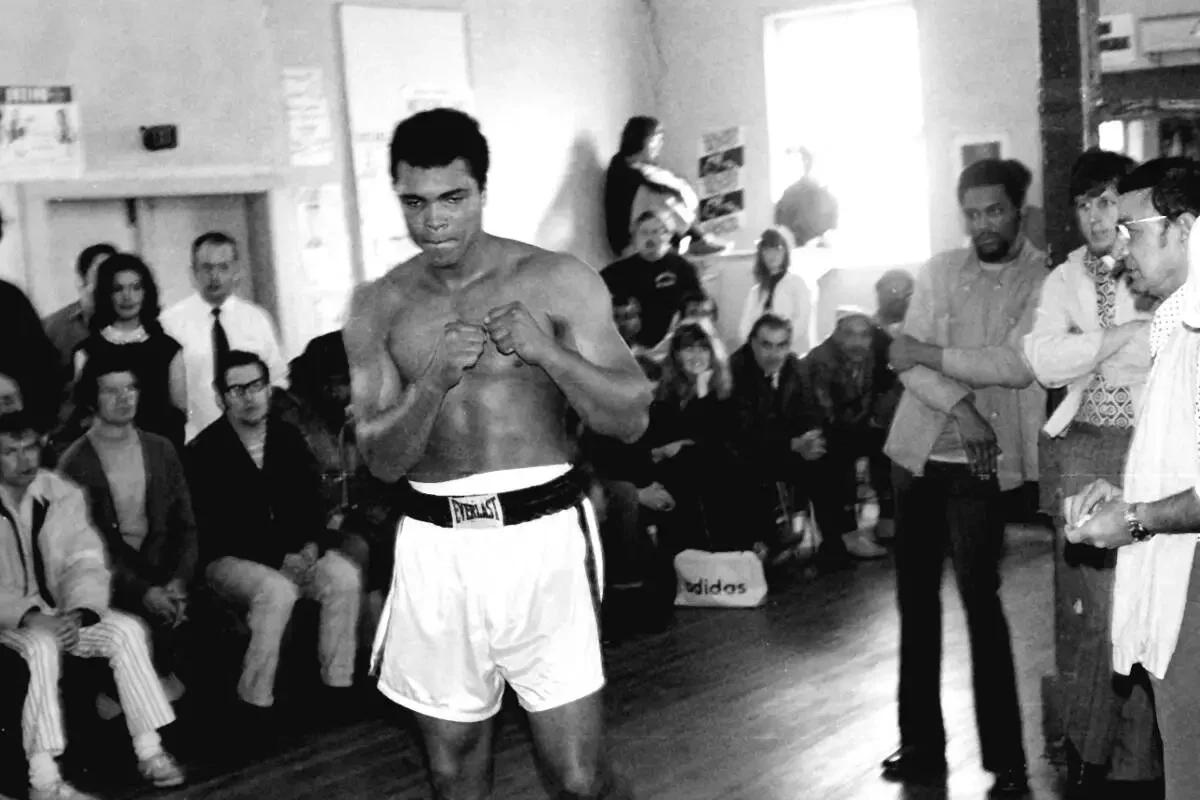 Boxer in boxing shorts standing with fists raised in a training or sparring session, surrounded by onlookers in an indoor gym or training space.