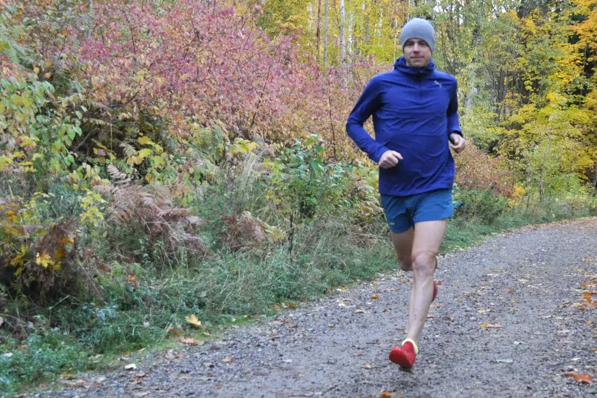A man running on a dirt trail in the woods during fall, wearing a blue jacket, blue shorts, a gray beanie, and red shoes, with colorful autumn leaves on the trees.