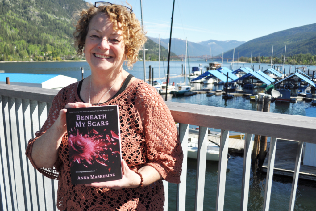 A woman with curly hair smiling and holding a book titled "Beneath My Scars" by Anna Maskerine, standing on a dock near a marina with boats and mountains in the background on a sunny day.