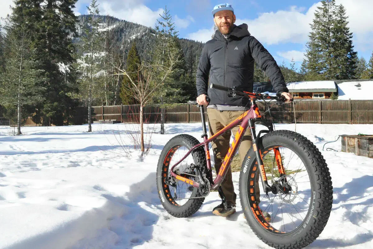 Man standing outdoors in snowy landscape with a fat tire mountain bike, trees, mountains, and a wooden fence in the background.