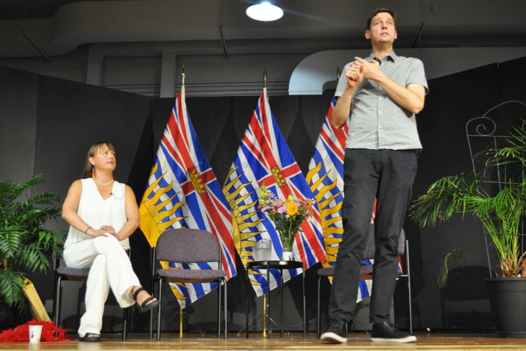 A man and a woman on stage in front of British Columbia flags. The man is standing and speaking, wearing a gray shirt and black pants. The woman is sitting on a chair, dressed in all white, listening attentively. There are potted plants and a small table with a bouquet of flowers between the flags.