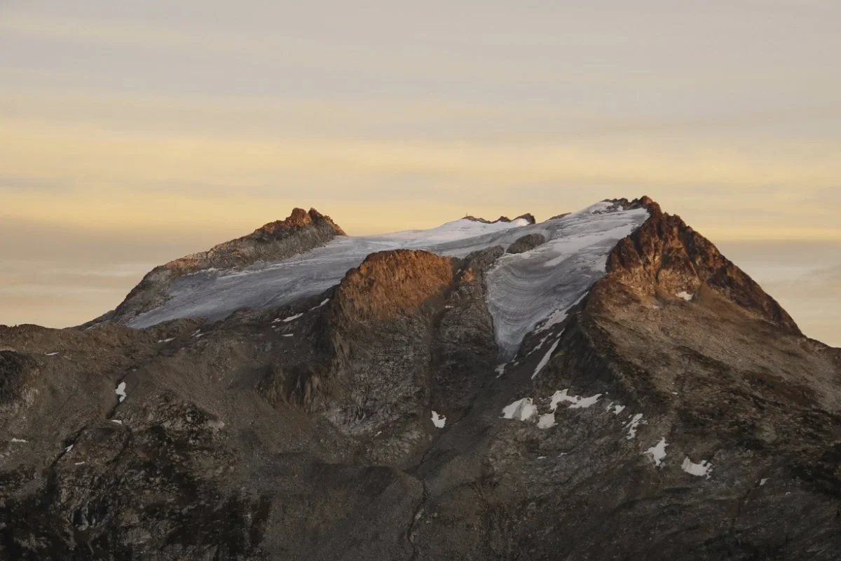 A mountain with rocky slopes and patches of snow, topped with ice and glaciers, under an orange-tinted sky at sunset.