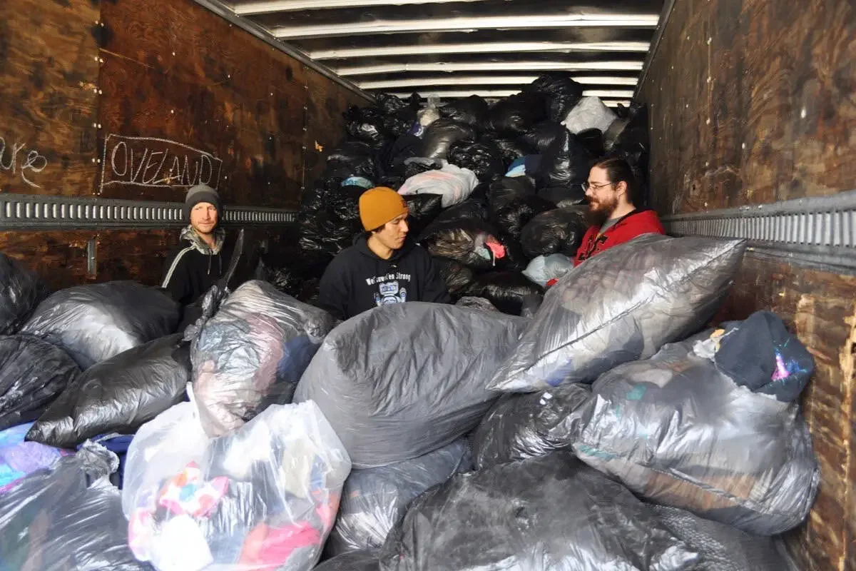 Three people standing inside a truck filled with black and gray garbage bags, with some bags spilling out and piled high in the background.