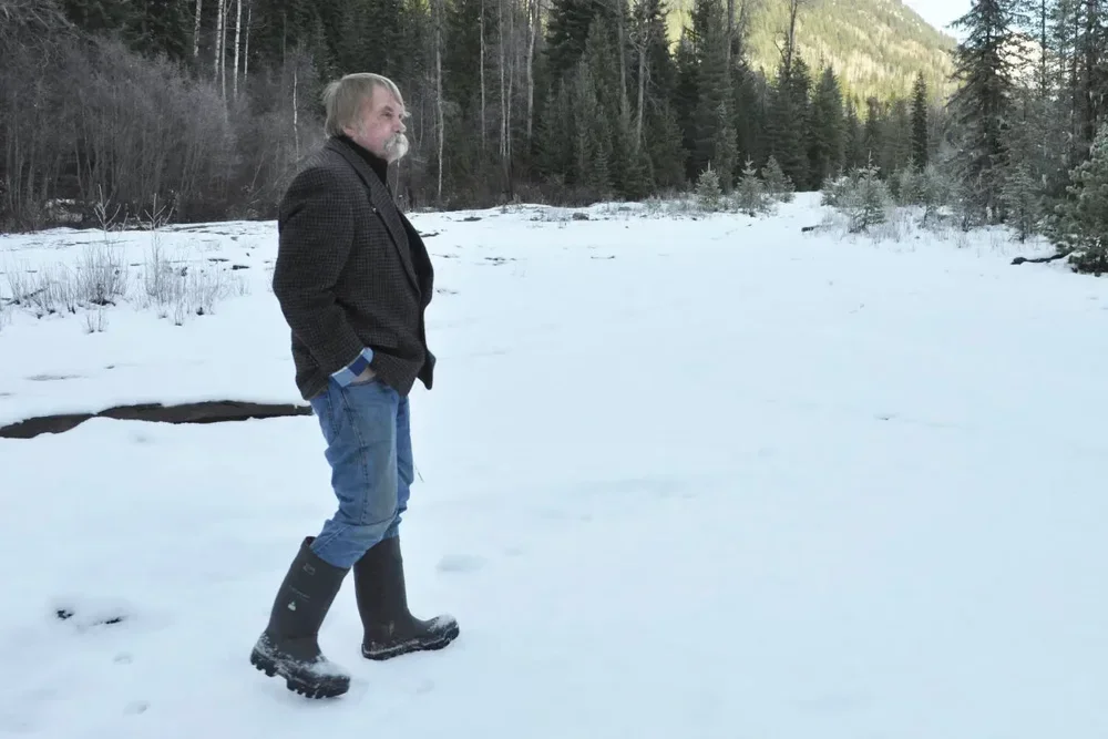 An older man standing in a snow-covered field with a forest and mountains in the background, wearing a black jacket, blue jeans, and black rubber boots.