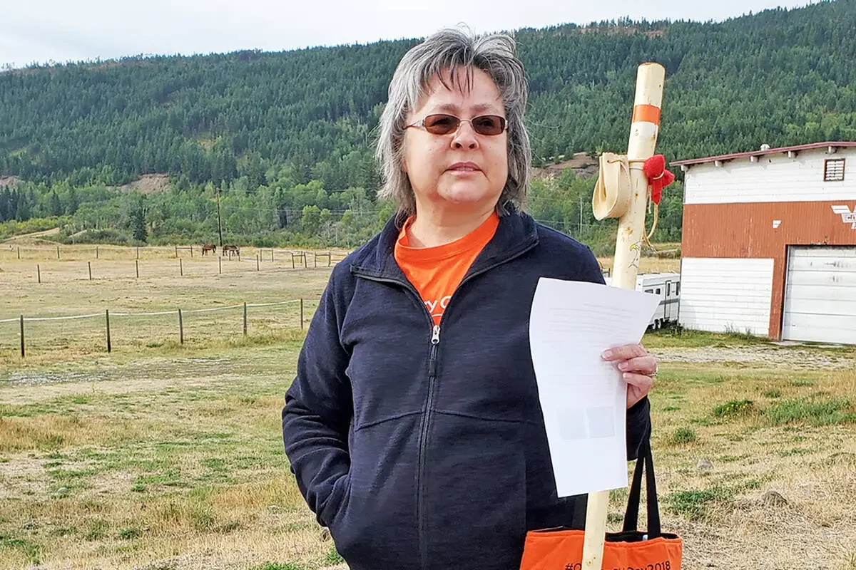 A woman with gray hair, sunglasses, and a serious expression stands outdoors in a rural area, holding a piece of paper and an orange tote bag. She is wearing a black jacket over an orange shirt. In the background, there is a large open field with a fence, a small building with a white and brown exterior, and green hills covered with trees.
