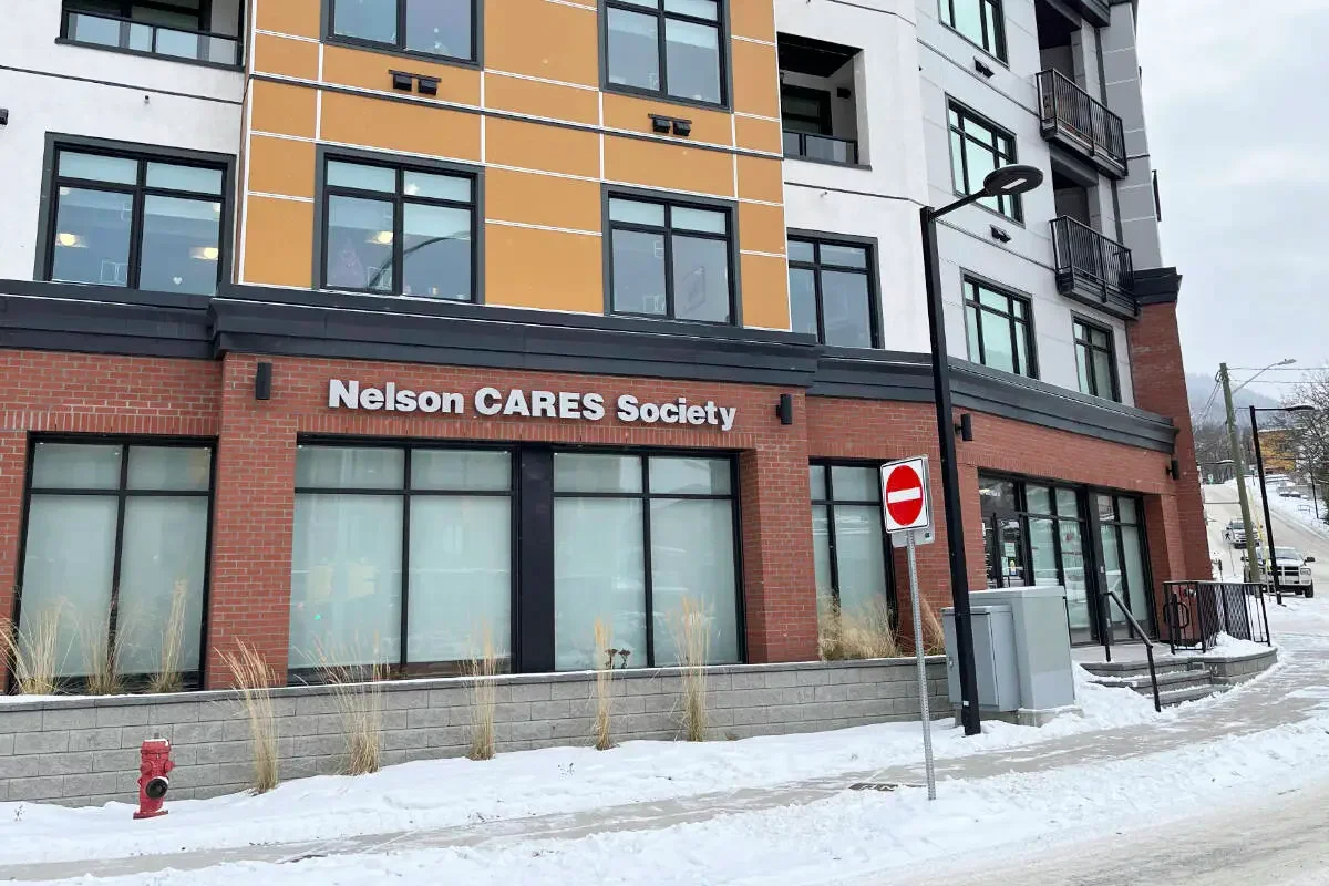 Exterior of a modern multi-story building with a sign that reads "Nelson CARES Society." Snow covers the sidewalk and nearby surroundings. There is a no entry sign and a street lamp in front of the building.