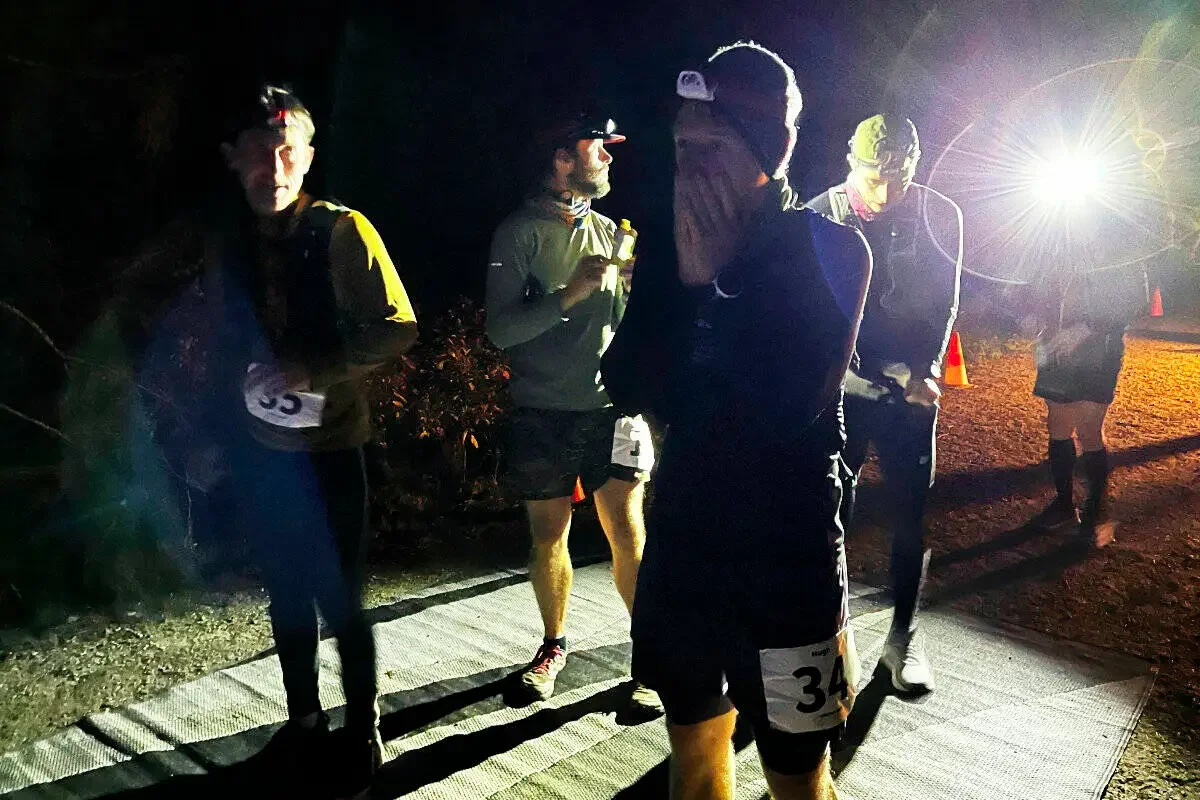 Group of runners at night preparing for a race on a lit trail, wearing athletic gear and numbered bibs.