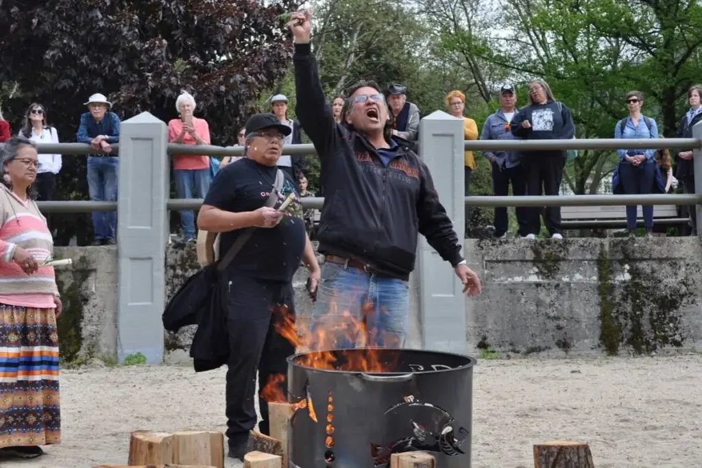 A man passionately protesting with a raised fist in front of a burning barrel at a public gathering, with onlookers watching behind a fence.