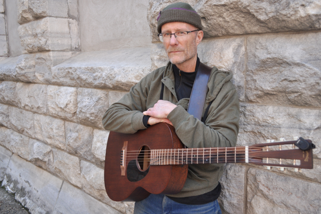 A man with glasses, a gray beanie, and casual clothing leaning against a stone wall while holding a guitar.